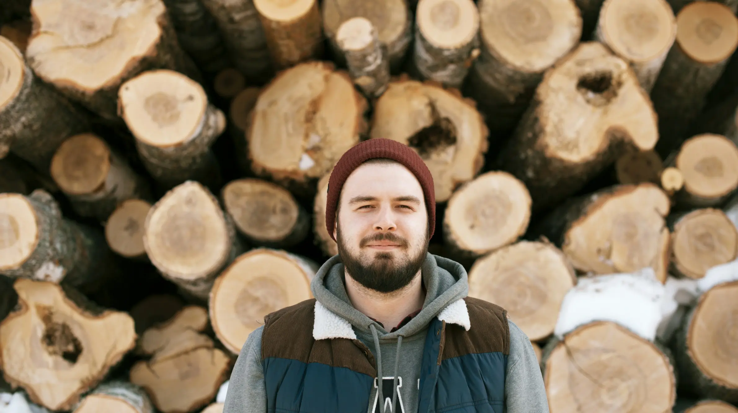 A bearded man wearing a maroon beanie, gray hoodie, and brown and blue jacket stands in front of a large stack of cut logs, with the cut ends of the logs facing the camera.